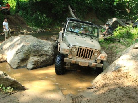 1996 Ford Bronco - A tan Jeep Wrangler drives through a muddy obstacle course with large rocks and trees.