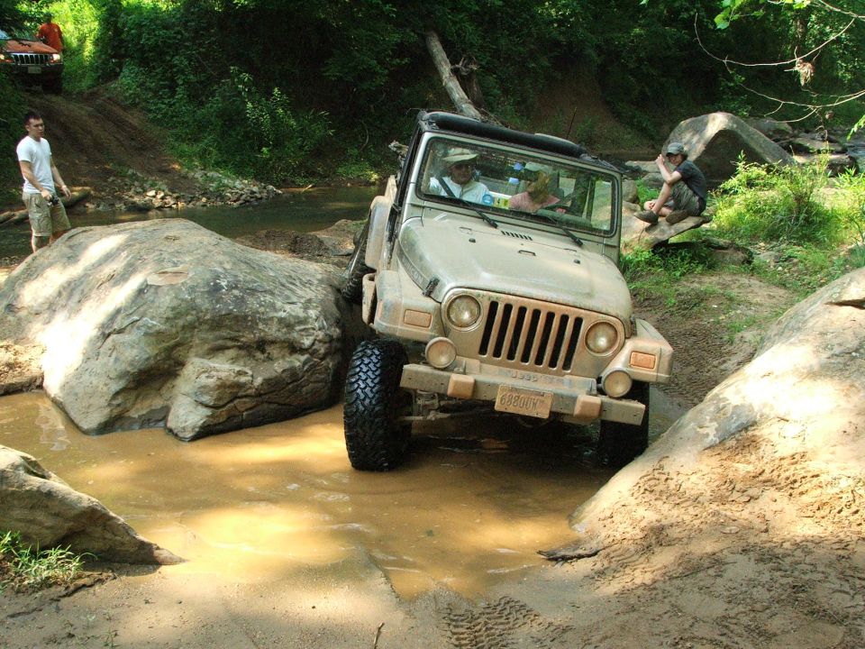 Tan Jeep Wrangler navigating a muddy stream with large rocks