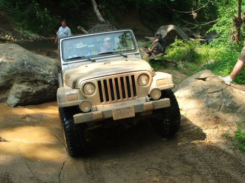 1996 Ford Bronco - A tan Jeep Wrangler is shown off-roading through a muddy creek bed with large rocks surrounding it.
