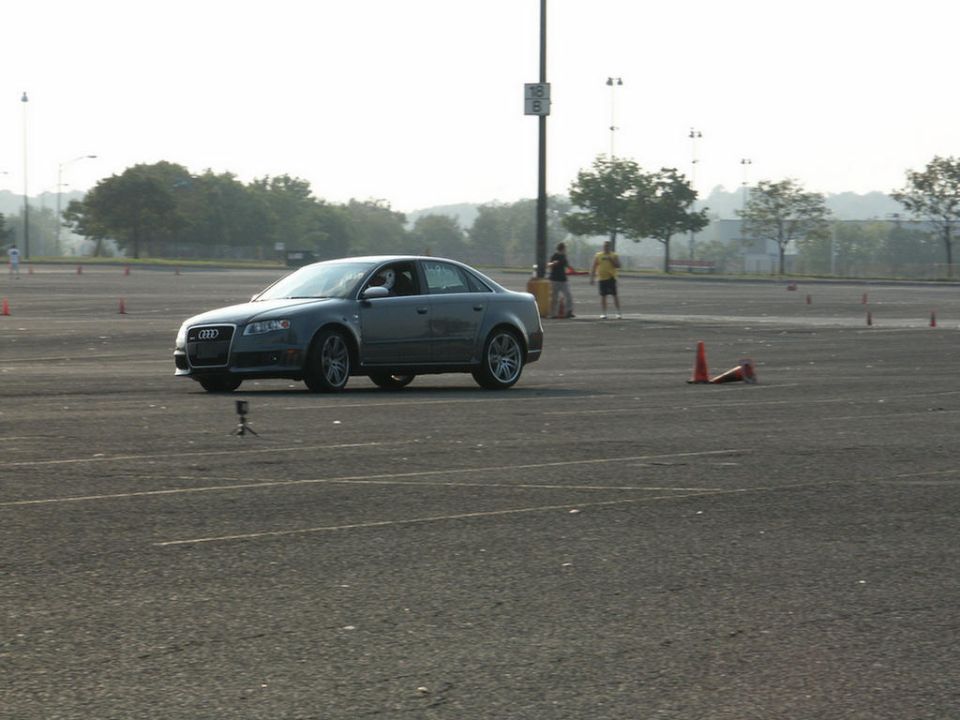 Gray Audi A4 sedan on a paved lot with cones, two people in background