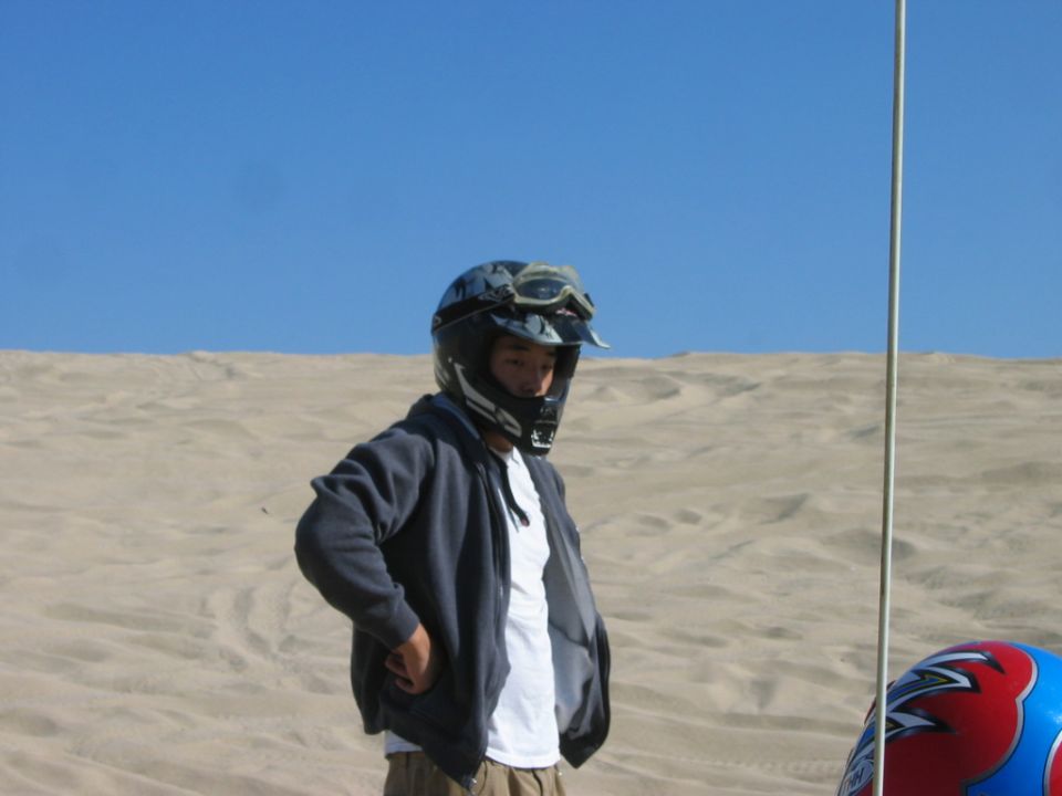 Person in dark helmet and hoodie standing on sand dunes under blue sky