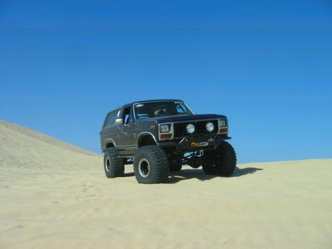 1985 Ford Bronco - This modified 1985 Ford Bronco sits on a sand dune under a clear blue sky, its lifted suspension and large off-road tires.