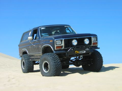 1985 Ford Bronco - Modified 1985 Ford Bronco with oversized tires and winch, parked on sand dunes under a clear blue sky.