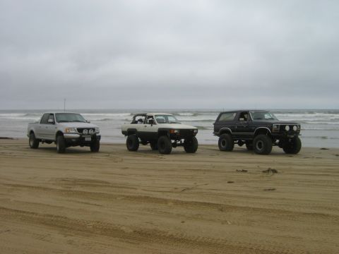 1985 Ford Bronco - Three modified trucks and SUVs are parked on a sandy beach under an overcast sky.