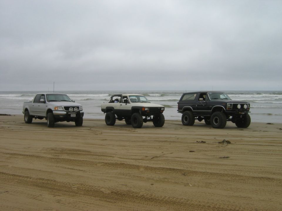 Three lifted 4x4 trucks and an SUV on oversized tires parked on a beach