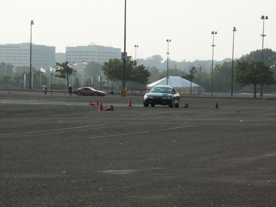 Black sedan on a paved lot with orange cones, red sports car in background