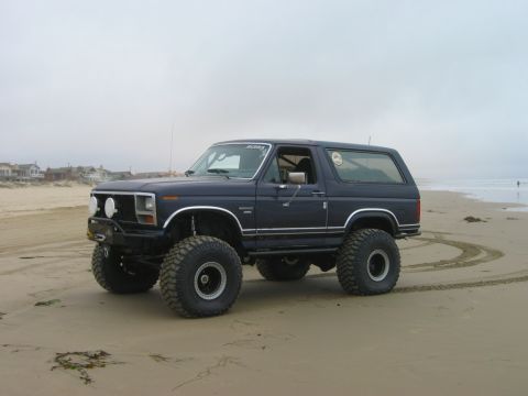 1985 Ford Bronco - A lifted 1985 Ford Bronco with large off-road tires parked on a sandy beach near the ocean.