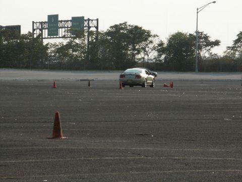 2001 Mercury Grand Marquis - A gold Ford Mustang navigates a course marked by traffic cones, with highway signage visible in the distance.