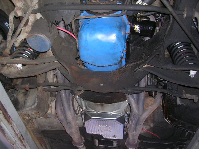 Underside view of a vehicle's engine, transmission, and suspension components. Shows a blue engine block, oil filter, exhaust headers, and springs