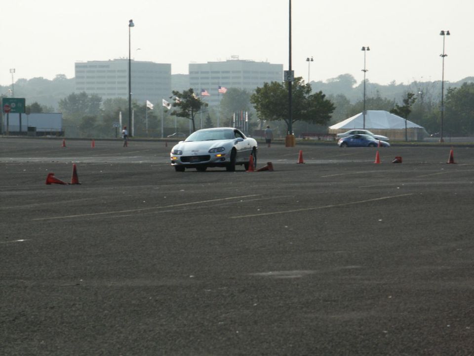 Silver Chevrolet Camaro driving on a paved lot with orange cones, buildings in background