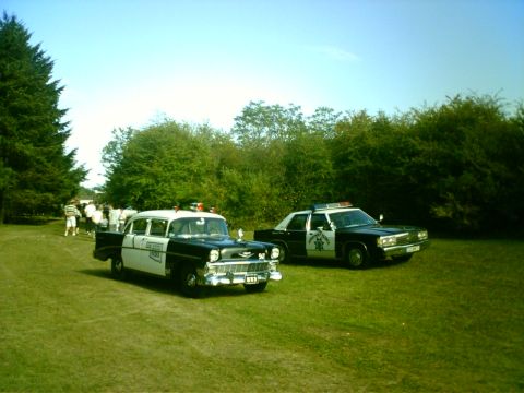 1991 Ford Crown Victoria - Two vintage police cars are parked on a grassy field under a clear blue sky.