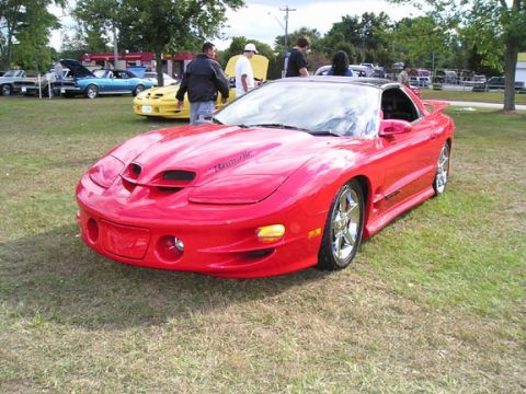 1994 Pontiac Trans Am - A red Pontiac Firebird is parked on a grassy field, with other vehicles and people visible in the background.