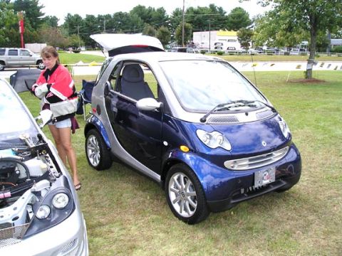 1994 Pontiac Trans Am - A blue and silver smart car is parked on grass with its trunk open. A woman stands nearby.
