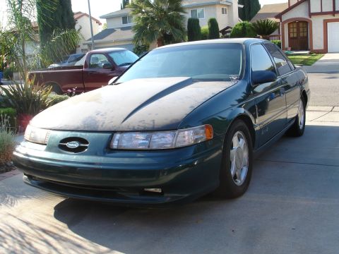 1993 Ford Taurus - Dark green 1993 Ford Taurus with a dusty hood sits in a driveway next to a red pickup truck.