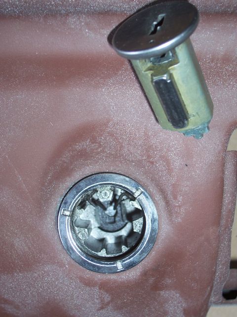 1978 Ford Bronco - Close-up of a removed door lock cylinder with its key slot and brass body, lying on a red-primered panel above the splined lock/handle bore…