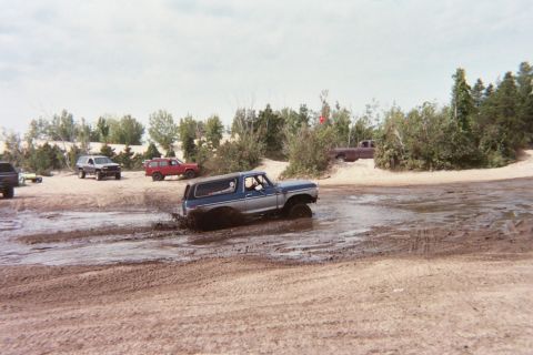 1979 Ford Bronco - Several off-road vehicles navigate a muddy terrain under a cloudy sky.