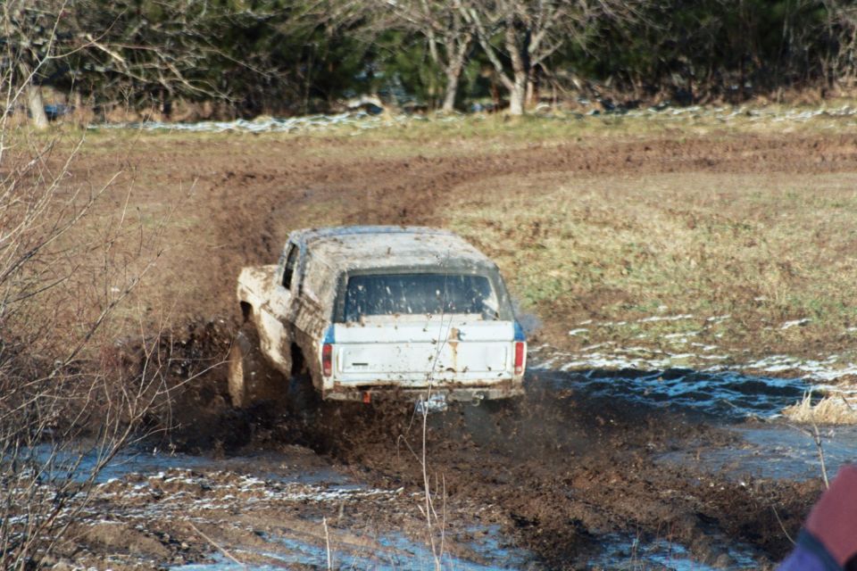 Mud-splattered white Ford Bronco driving through a muddy track, rear view