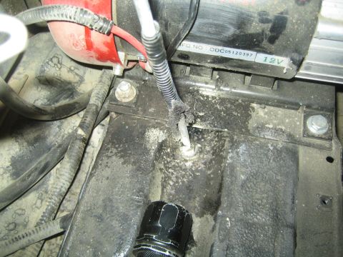 1990 Ford Bronco - engine bay view with a corroded battery hold-down rod and bolt, surrounding wiring, a black socket and a red component, labeled 12V.