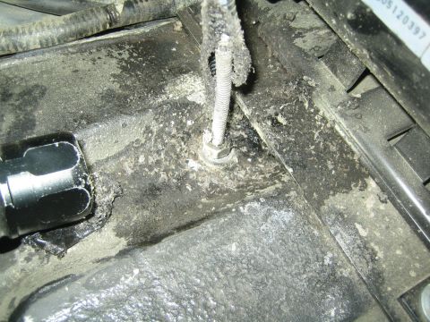 1990 Ford Bronco - close-up of a corroded threaded hold-down bolt rising from a dirty battery tray, with a black socket and grime around it.
