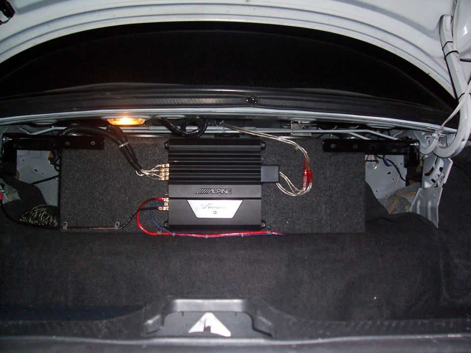 Car trunk interior showing an Alpine V-Power amplifier mounted on a carpeted enclosure