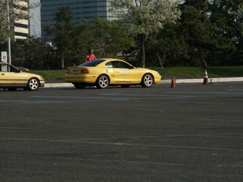 2001 Mercury Grand Marquis - A yellow Ford Mustang is parked on a paved lot with cones, next to another yellow car.
