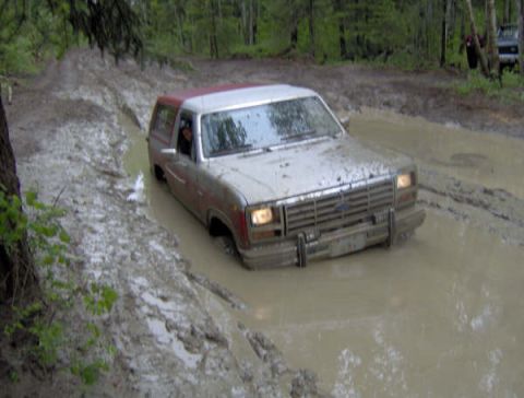 1989 Ford Bronco - A silver Ford Bronco navigates a deep mud pit on a wooded trail.