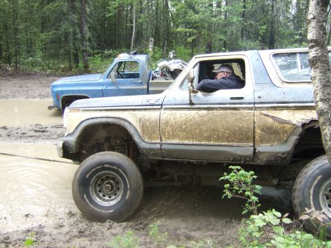 1989 Ford Bronco - A heavily mud-caked 1978 Ford Bronco is being pulled out of a muddy ditch by a blue pickup truck in a wooded area.