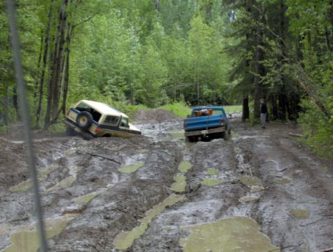 1989 Ford Bronco - Two off-road vehicles navigate a challenging muddy trail through a forest.