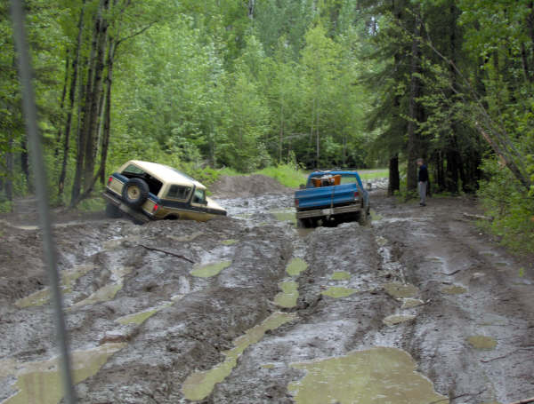 Two off-road vehicles stuck in deep mud on a wooded trail. One is a tan SUV, the other a blue pickup truck