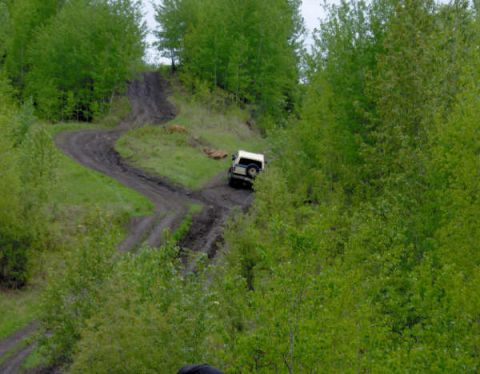 1989 Ford Bronco - A white 1989 Ford Bronco navigates a challenging muddy off-road trail surrounded by lush green trees.
