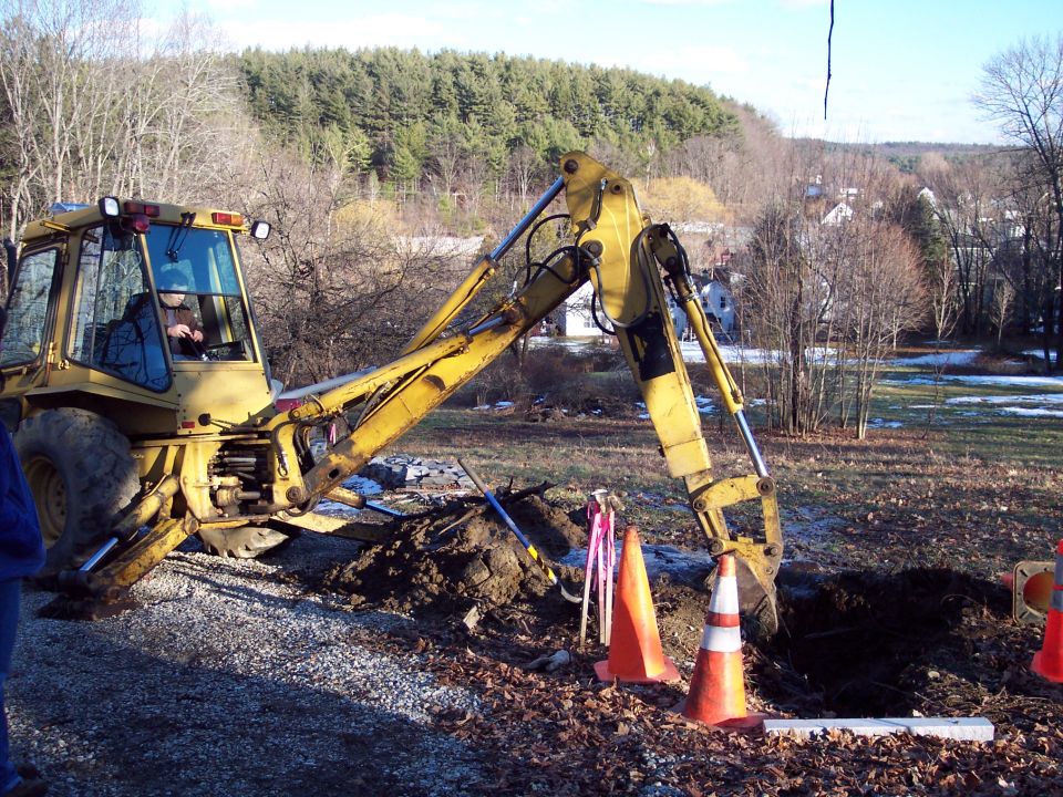 Yellow backhoe loader digging a trench with orange cones nearby. Trees and houses in background