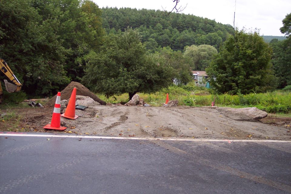Construction scene with two orange traffic cones, a pile of dirt, and a large rock on a dirt road next to a paved road