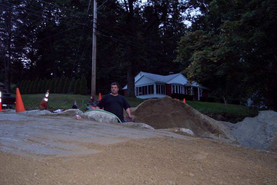 Man standing at a construction site with piles of dirt and sand, traffic cones, and a house in the background