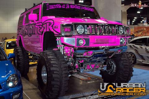 1978 Ford Bronco - Three-quarter front view of a hot pink lifted Hummer H2 on an indoor car-show floor, riding on massive mud-terrain tires with polished whee…