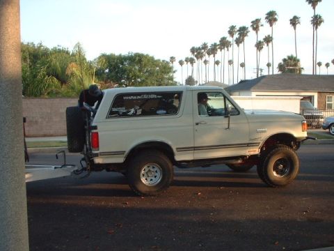 1966 Ford Bronco - A white full-size Ford Bronco with a lift kit, oversized mud tires, and a rear-mounted spare tire, shown in side profile parked on a street…