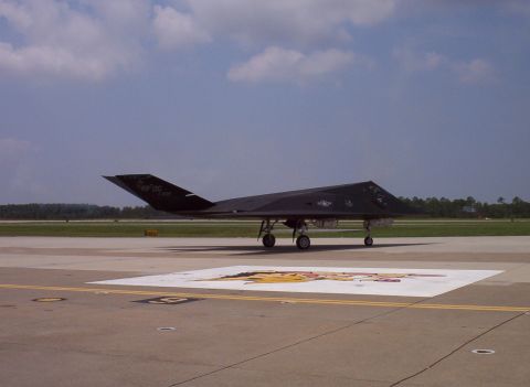 2003 Ford Crown Victoria - An F-117 Nighthawk stealth fighter jet is parked on an airfield tarmac under a partly cloudy sky.