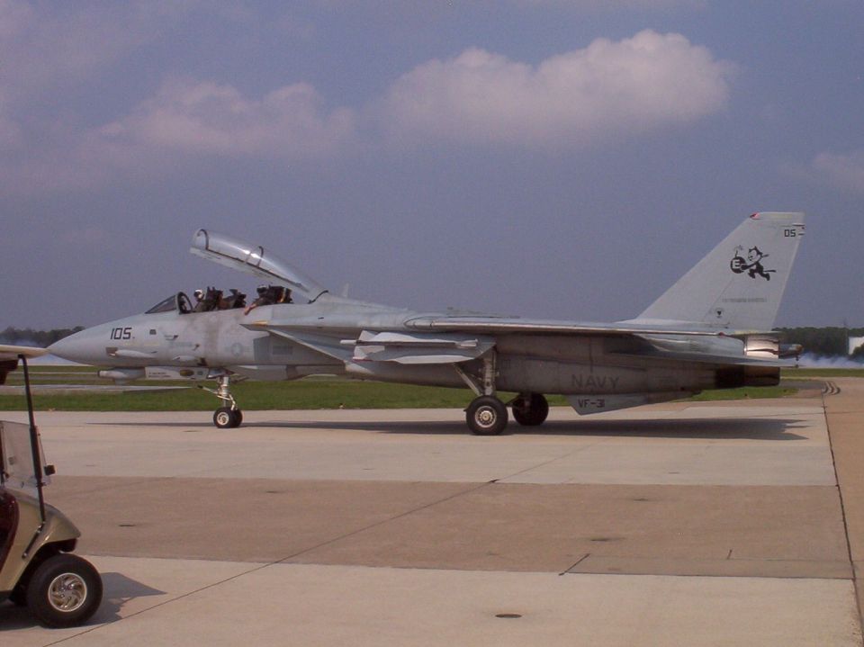 F-14 Tomcat fighter jet on tarmac, canopy open. Golf cart in foreground