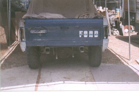 1966 Ford Bronco - Rear view of a blue early Ford Bronco with a tan soft top draped over the body, showing the weathered tailgate with chrome FORD block lette…