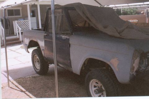 1966 Ford Bronco - Side view of an early Ford Bronco project in mixed primer and faded blue paint, doors off and a tarp draped over the roll bar, parked under…