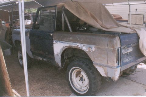 1966 Ford Bronco - Rear three-quarter view of an early Ford Bronco project under a carport, doors removed and a tan tarp draped over the exposed roll bar and…