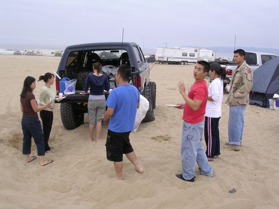 People gathered around a lifted SUV on a sandy beach, with RVs and other vehicles in the background