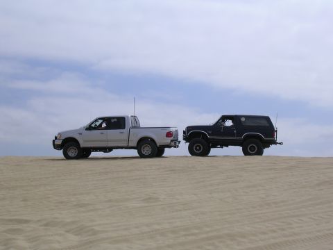 1985 Toyota 4Runner - Two trucks, a dark blue SUV and a silver pickup, are parked side-by-side on a sandy terrain with a cloudy sky above.