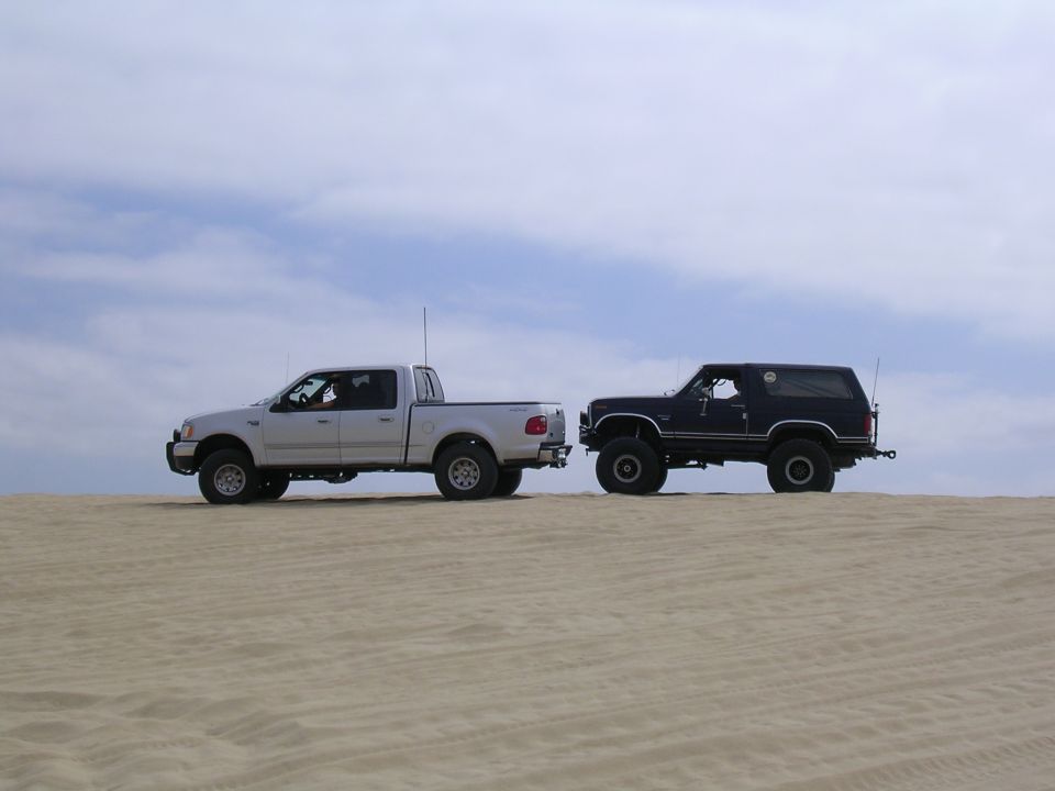 Dark blue SUV and silver pickup truck parked on sand dunes under a cloudy sky