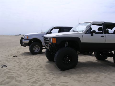 1985 Toyota 4Runner - Two modified SUVs sit on a sandy terrain under an overcast sky.