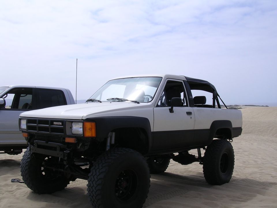 White and black Toyota 4Runner with large tires, lifted suspension, and a roll bar. The vehicle is on a sandy terrain