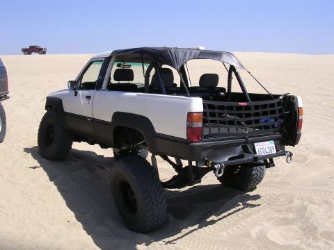 1985 Toyota 4Runner - Modified Toyota pickup truck with a soft top and off-road tires parked on sand, with another vehicle in the distance.