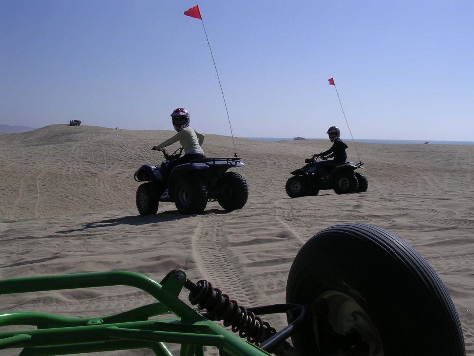 Green roll cage and tire in foreground, two quad bikes with red flags on sand dunes under a clear blue sky