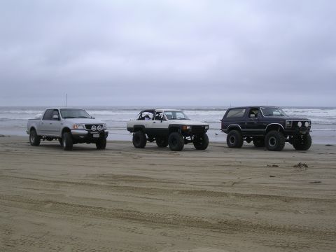 1985 Toyota 4Runner - Three modified off-road vehicles are parked on a beach under an overcast sky.