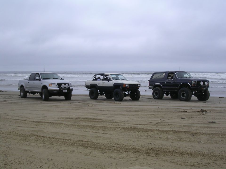 Silver pickup, white Toyota 4Runner, and dark blue SUV parked on a sandy beach with the ocean in the background