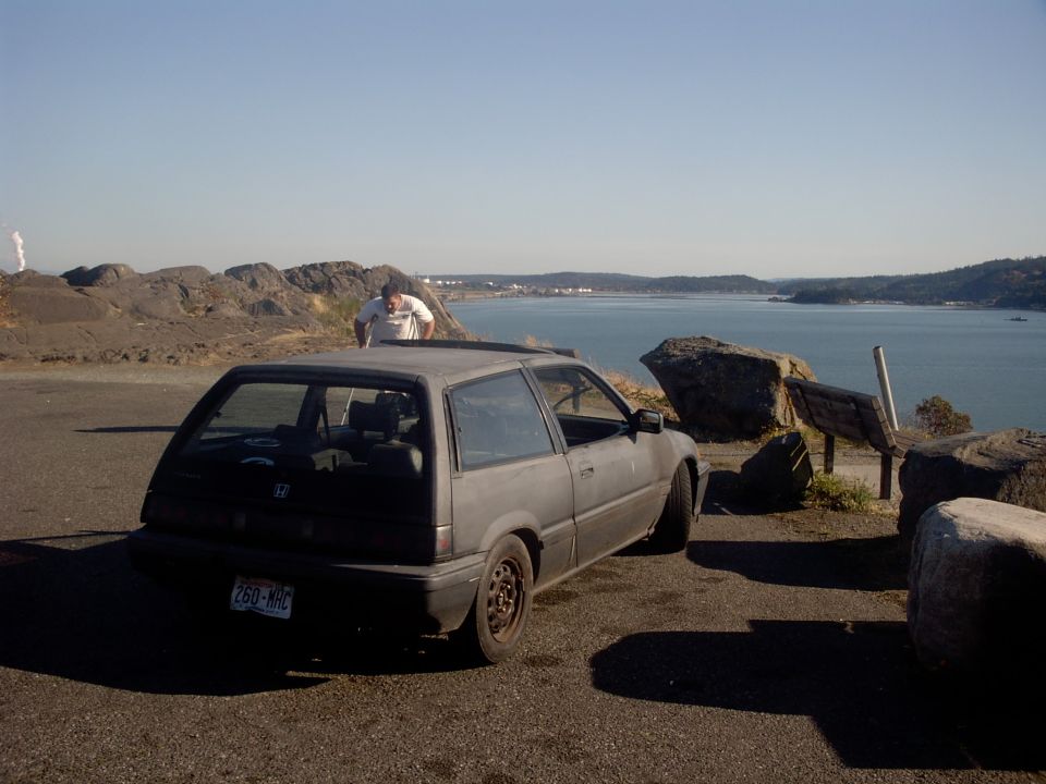 Primer-black Honda Civic hatchback on steel wheels at a waterfront overlook, 3/4 rear view